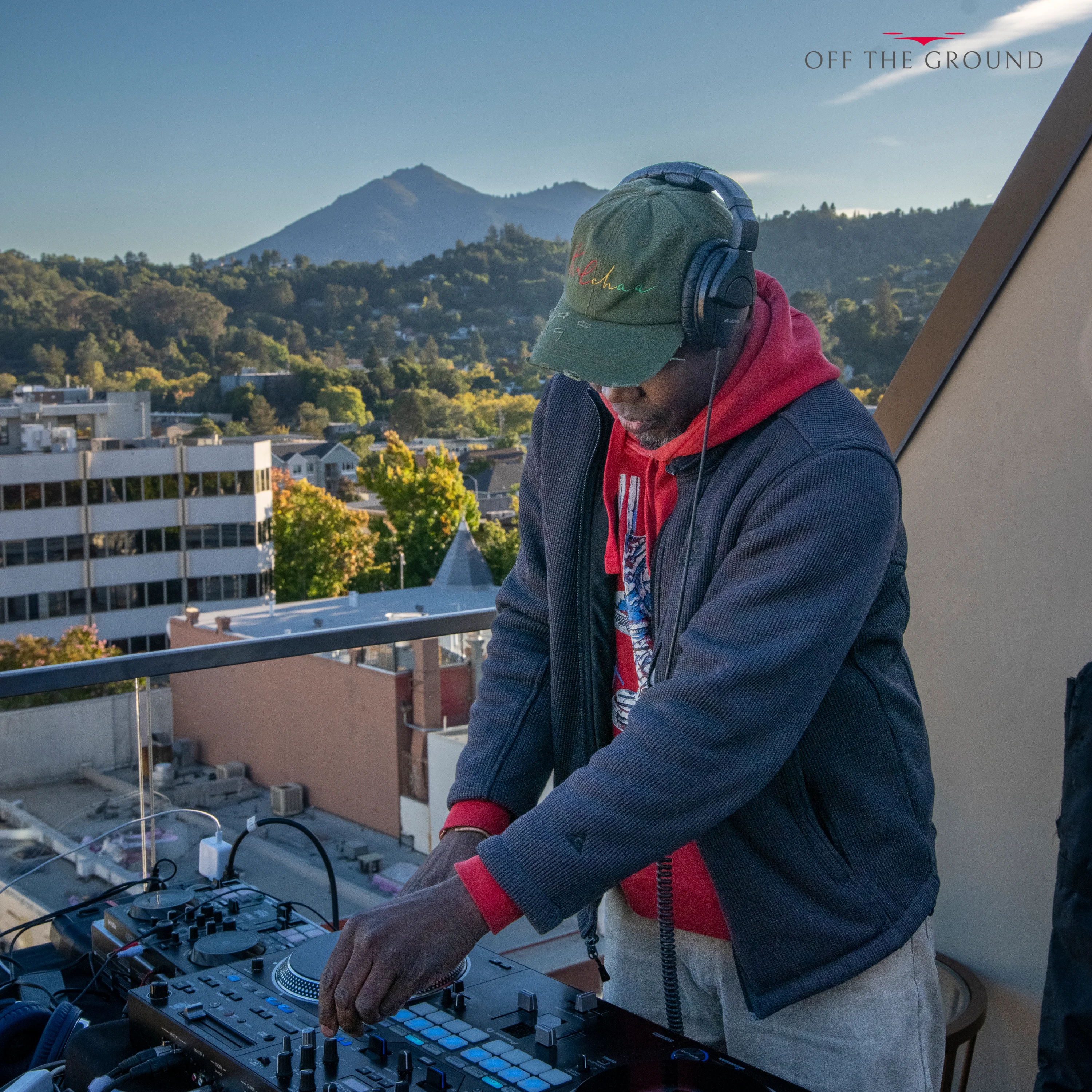 A DJ in a cap and red scarf working the decks on the rooftop with a mountain behind