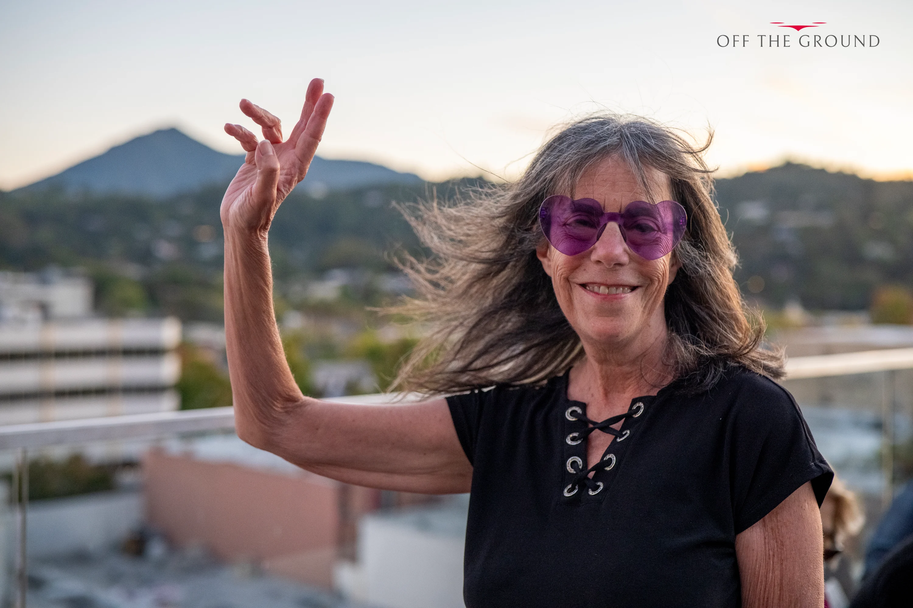 A guest in purple sunglasses raising one hand and smiling on the rooftop, hills in the background